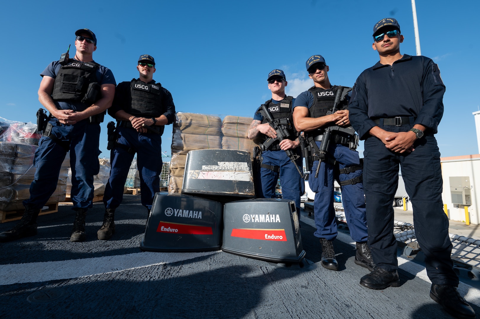 Law enforcement crew members from USCGC Valiant (WMEC 621) stand in front of interdicted narcotics and engine covers at Base Miami Beach, Florida, Mar. 6, 2025. The Valiant's crew secured the illegal drugs after six interdictions in the international waters of the Caribbean. (U.S. Coast Guard photo by Petty Officer 3rd Class Nicholas Strasburg)