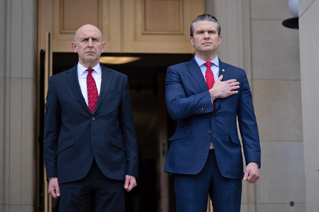 Two men in business suits are standing next to each other. The man on the right is holding his hand over his heart.