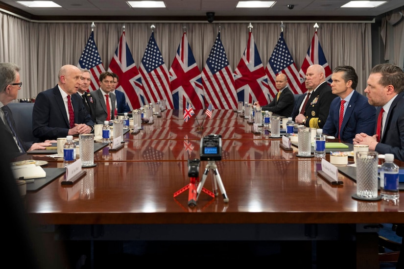 A group of nearly one dozen men in business suits are seated on opposite ends of a large wooden table. Flags of the U.S. and United Kingdom are on the wall behind them.