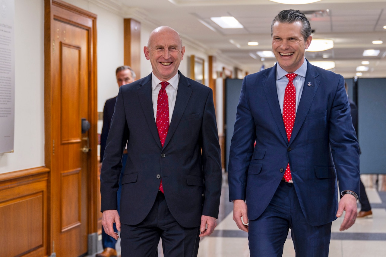 Two men in business suits are smiling while walking down a hallway.