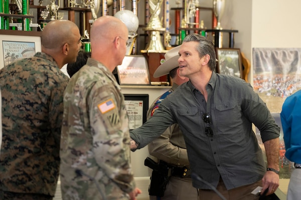 The defense secretary shakes hands with two service members in a room with trophies in the background.