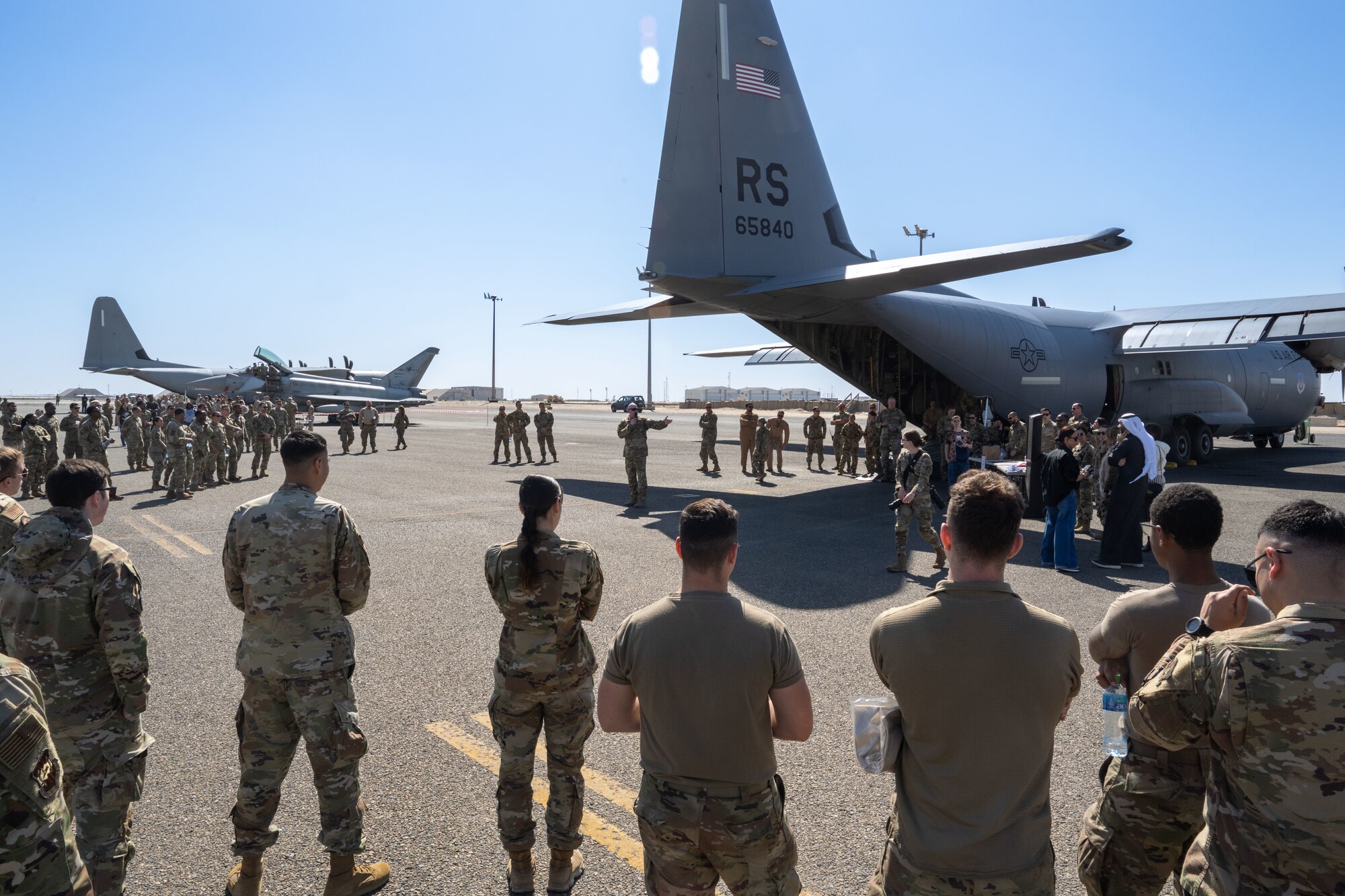 Service members in uniform listen to a leader speak in front of a plane.