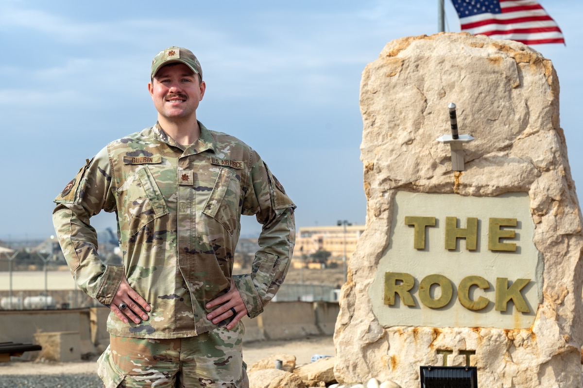 A man in uniform poses for a photo next to The Rock monument with a U.S. flag flowing behind the monument.