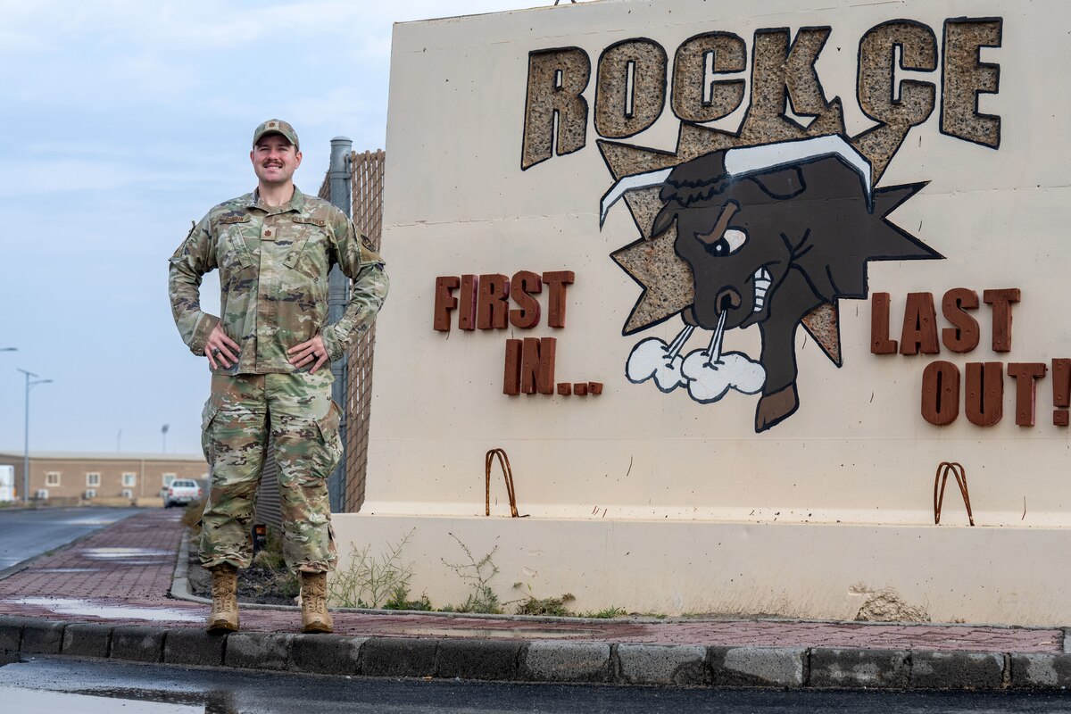 A man in uniform stands next to a T-Wall with a graphic of his squadron painted on the wall.