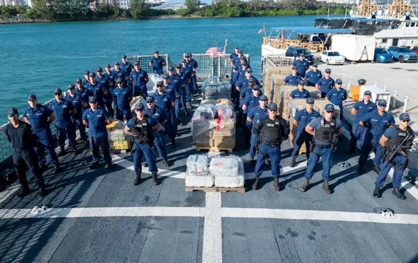 Crew members from USCGC Valiant (WMEC 621) stand at parade rest in front of interdicted narcotics at Base Miami Beach, Florida, Mar. 6, 2025. The Valiant's crew secured the illegal drugs from six interdictions in the international waters of the Caribbean. (U.S. Coast Guard photo by Petty Officer 3rd Class Nicholas Strasburg)