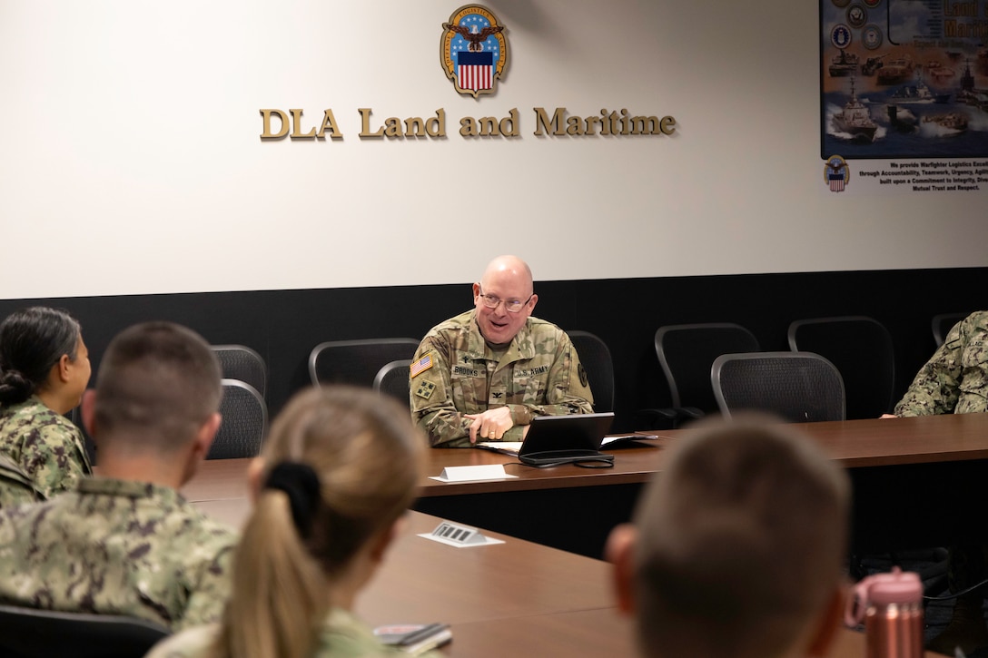 A man in a military uniform sits at the head of a table in a meeting with several others in military uniform.