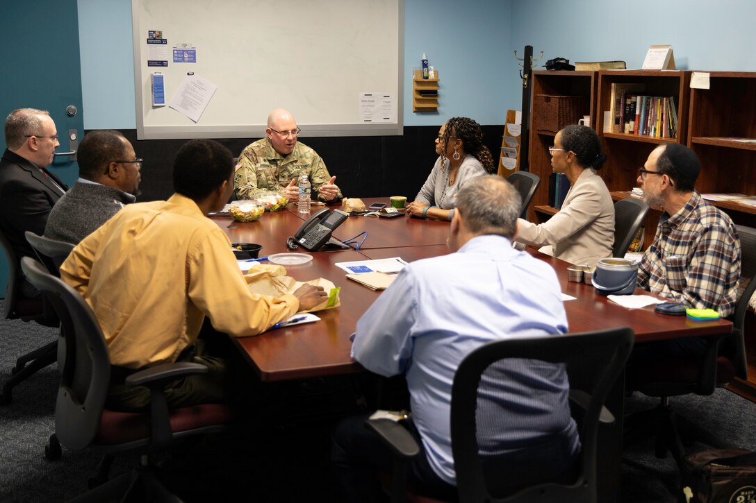 A man in a military uniform sits at the head of a table as he meets with a group of seven people.