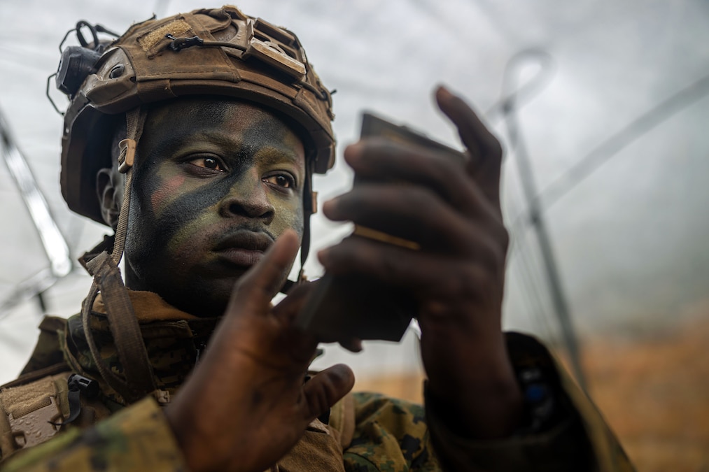 A Marine applies camouflage while wearing combat gear and holding a case.