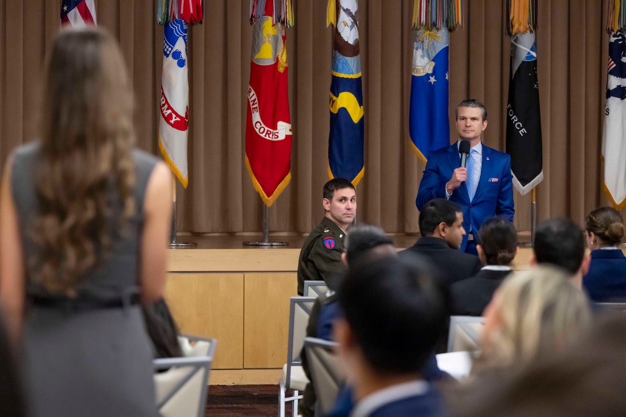 A man stands at the front of a room while a young lady stands facing him to ask a question.