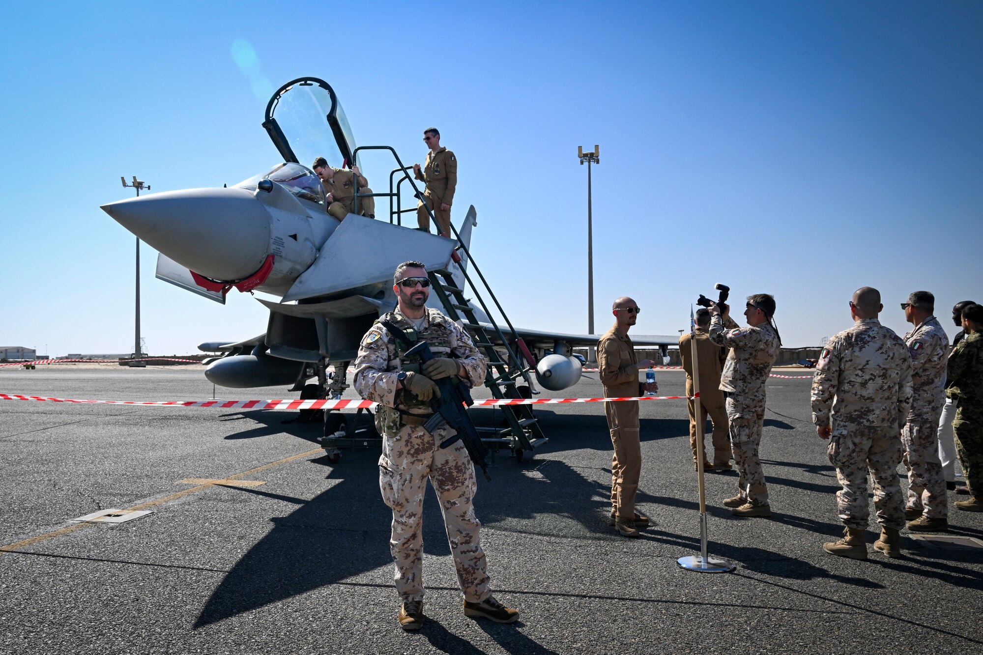 A man in uniform stands in front of a fighter jet with a rifle.