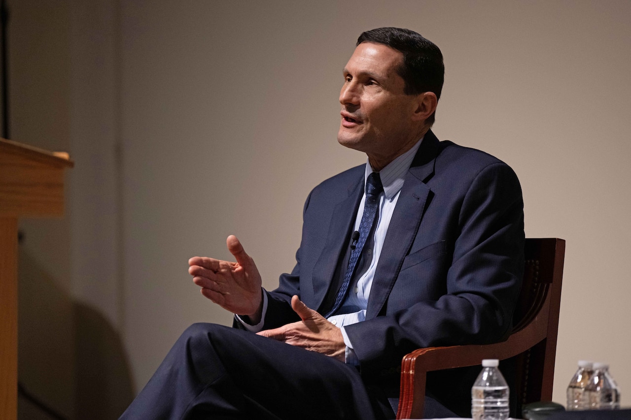 A man wearing a business suit is speaking while sitting in a chair.
