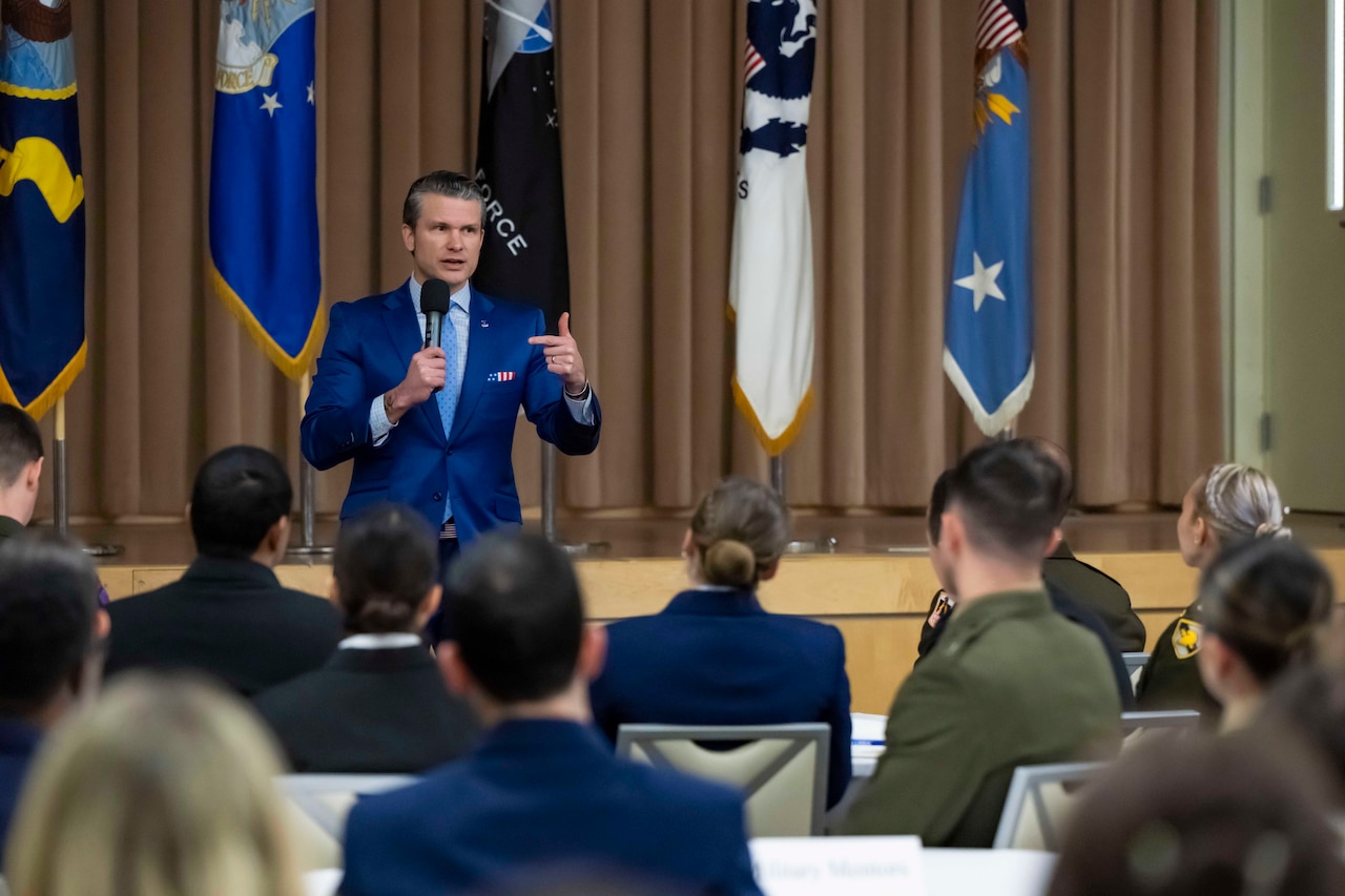 Defense Secretary Pete Hegseth speaks to an audience while holding a mic with a stage and flags in the background.