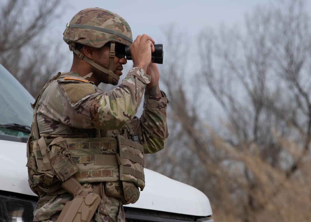 A soldier uses binoculars while standing next to a partially visible white vehicle with trees in the background.