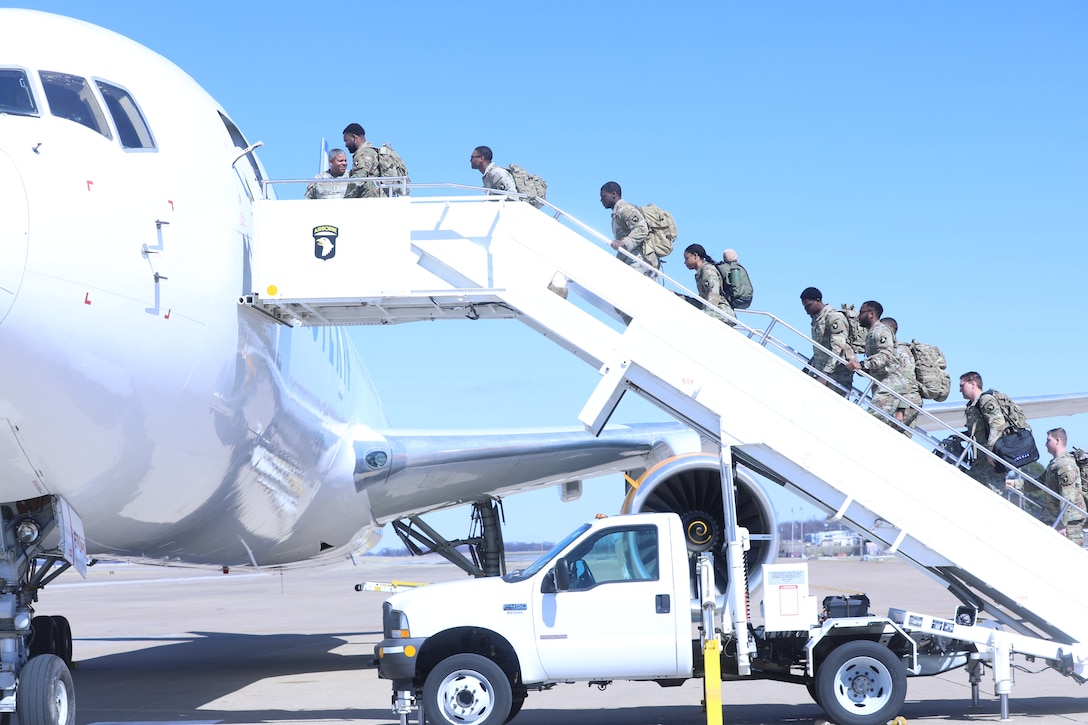 Soldiers carrying backpacks walk up steps onto an airplane against a blue sky.