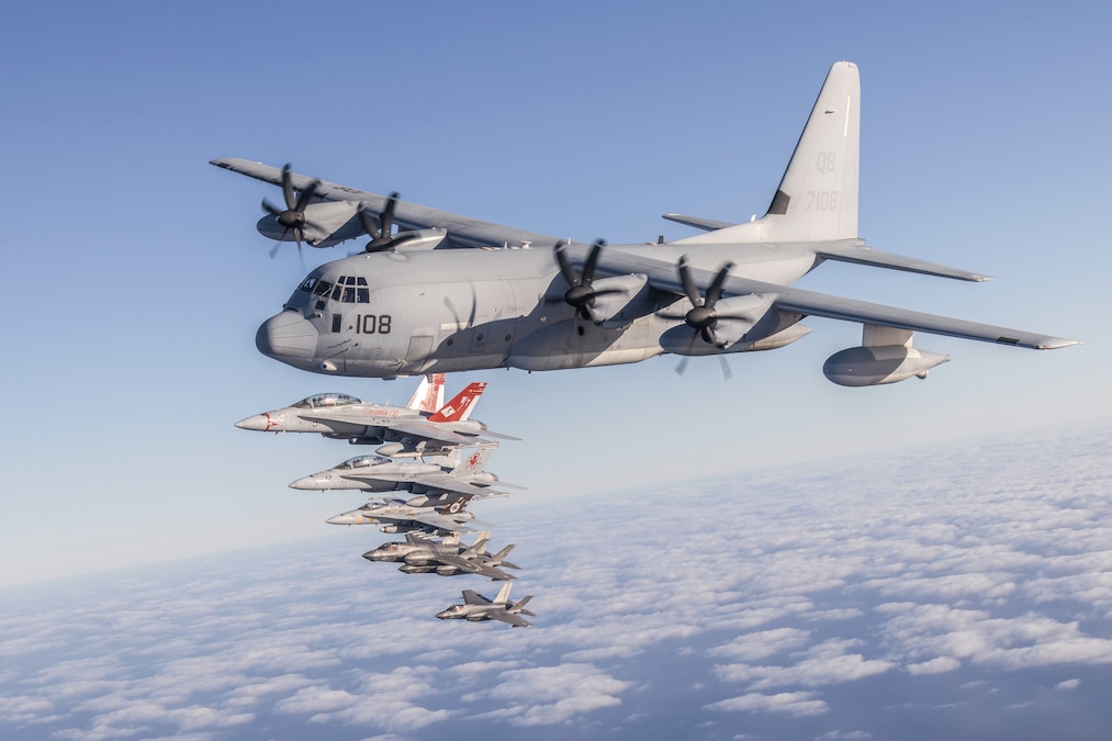 A large aircraft flies in formation with a group of smaller aircraft above clouds during the day.