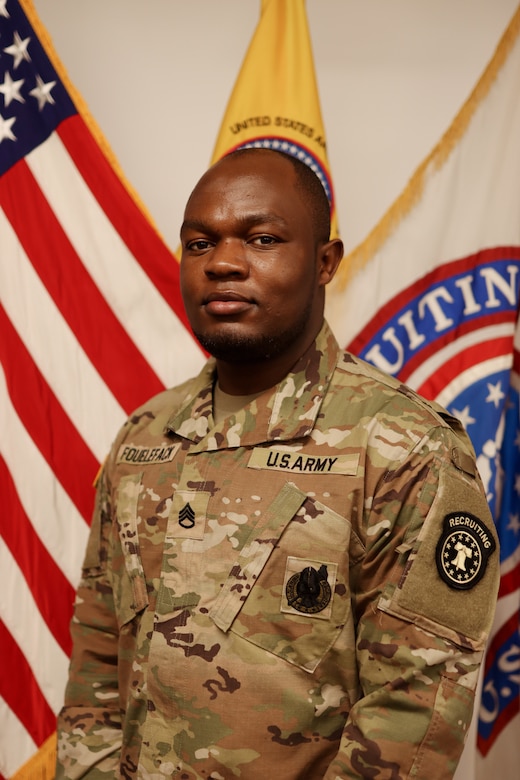 Male U.S. Army Soldier poses in uniform in front of the American flag and USAREC flag