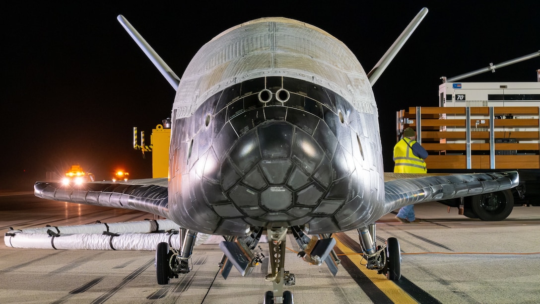 A rounded vehicle with wings sits on a flight line lit up in the dark.
