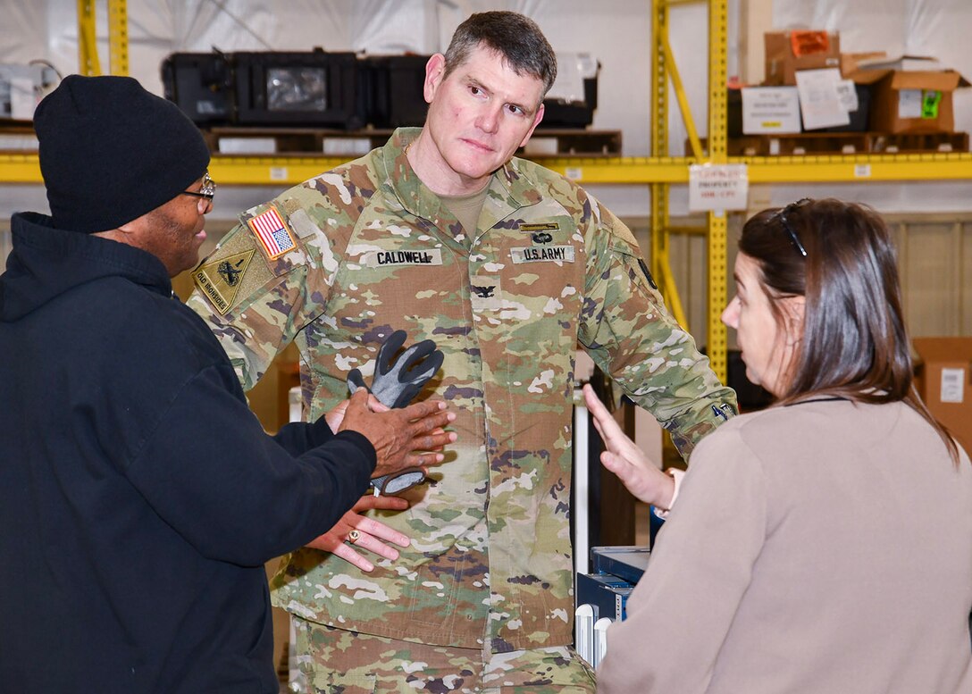 Two men, one dressed in the uniform of an Army Col. and a woman stand together in deep conversation. They are standing in a warehouse.