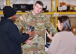 Two men, one dressed in the uniform of an Army Col. and a woman stand together in deep conversation. They are standing in a warehouse.