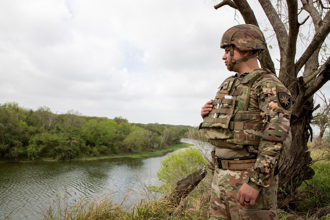 A soldier look out to a body of water while standing in a forest.