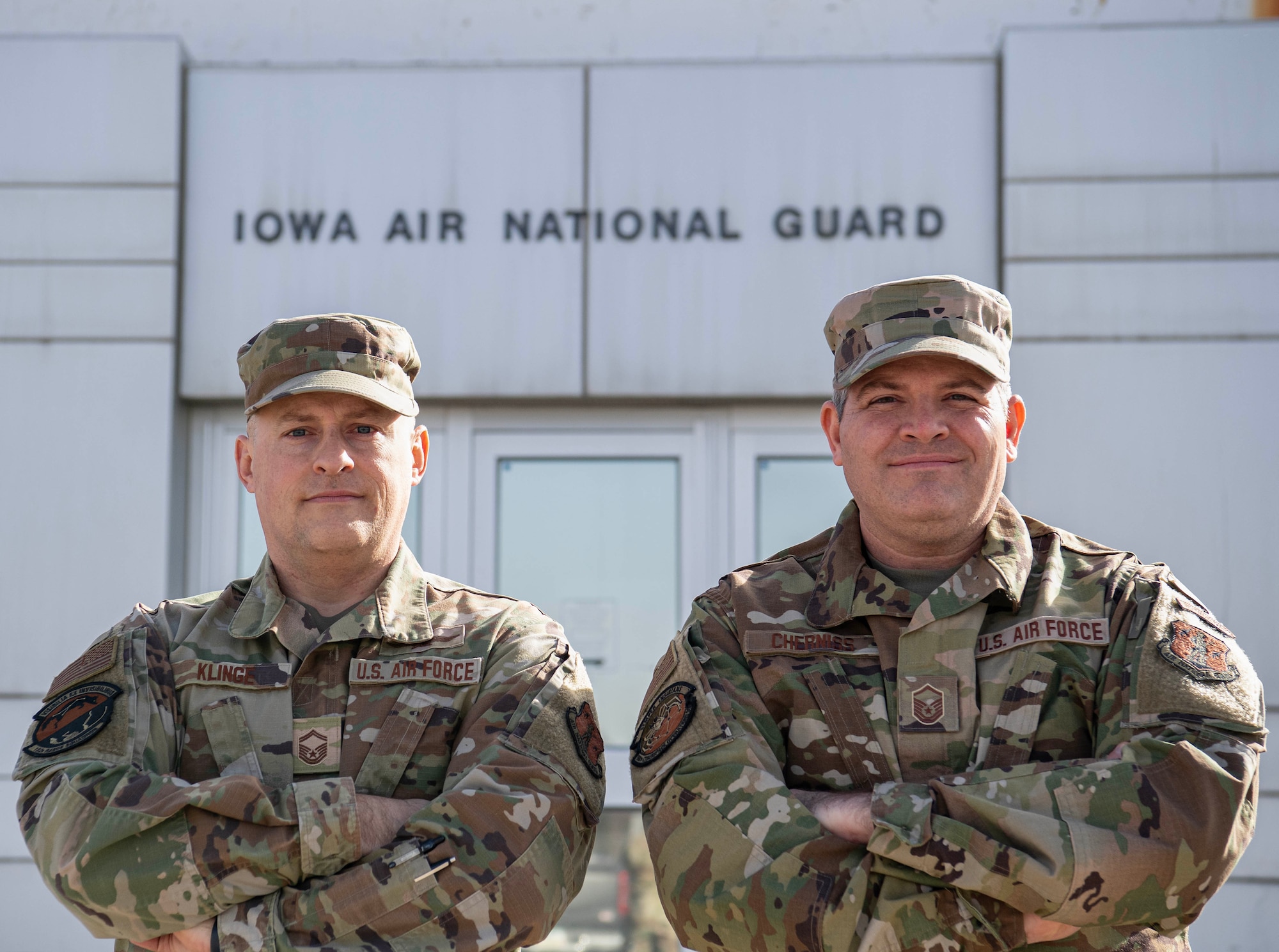 Two uniformed male Airmen stand with crossed arms posing for a photo with a building in the background that says Iowa Air National Guard