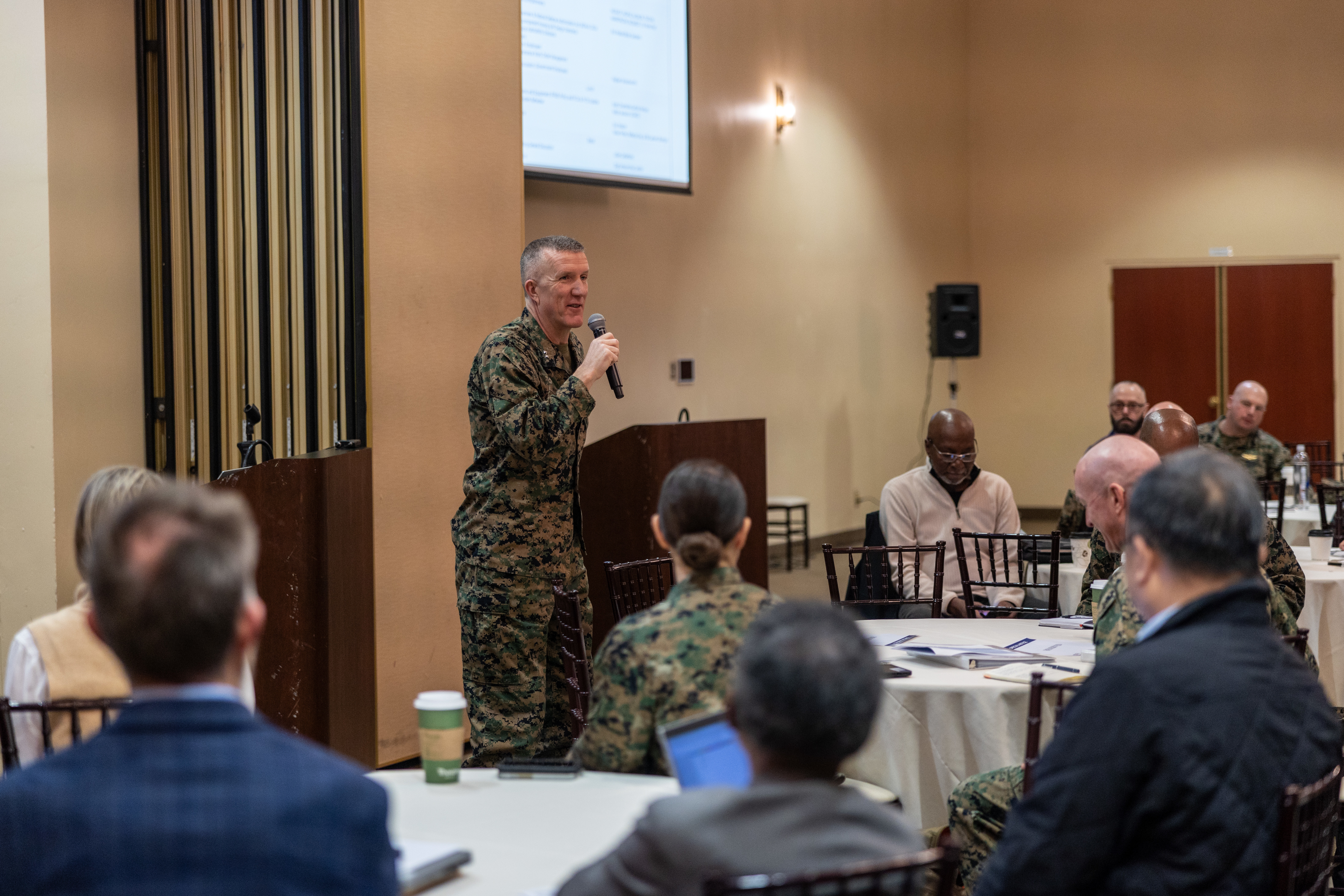 MajGen Maxwell speaking into a microphone for a group of people sitting at tables.