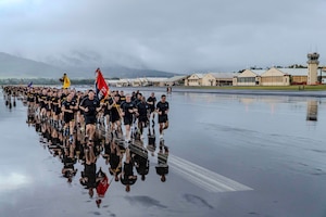 A large group of soldiers runs in rows of four on a wet flight line under cloudy skies.