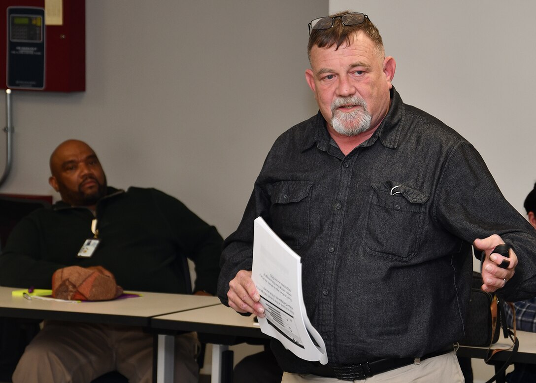 A man in a black shirt talks with a person seated behind him.