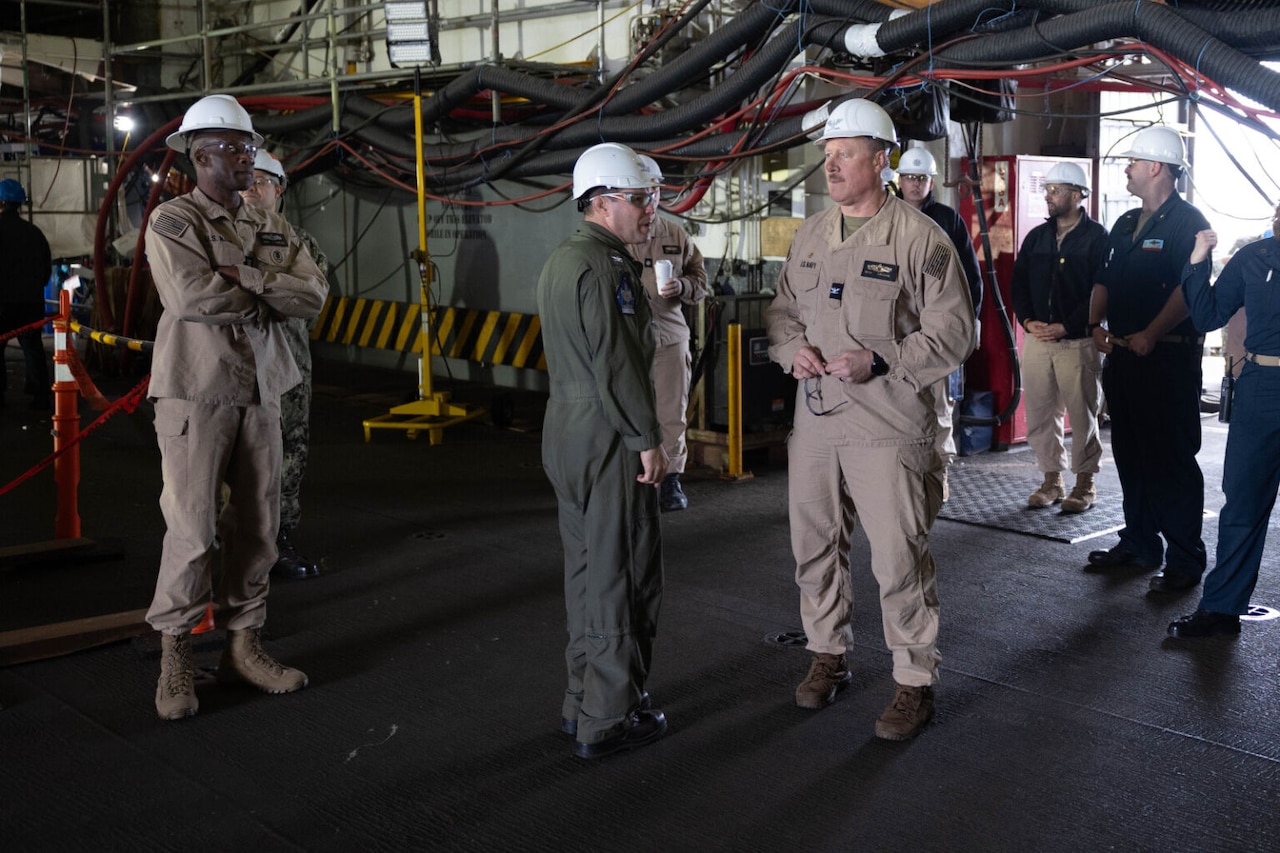 Sailors wearing hard hats talk while aboard a ship.