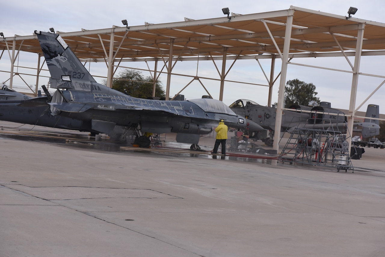 A man washes an airplane parked on a tarmac under a steel structure.