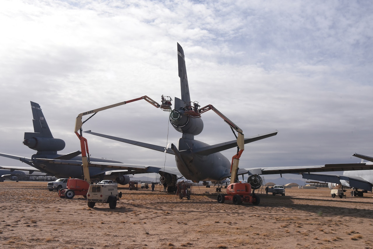 Workers raised high by crane equipment work on the tail of an airplane parked in dirt.