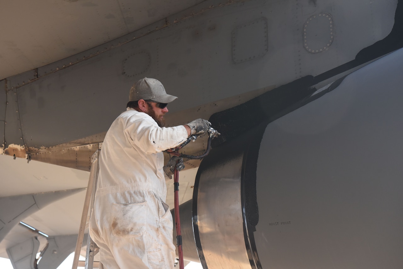 A worker in white coveralls sprays a coating onto an airplane with a hose.