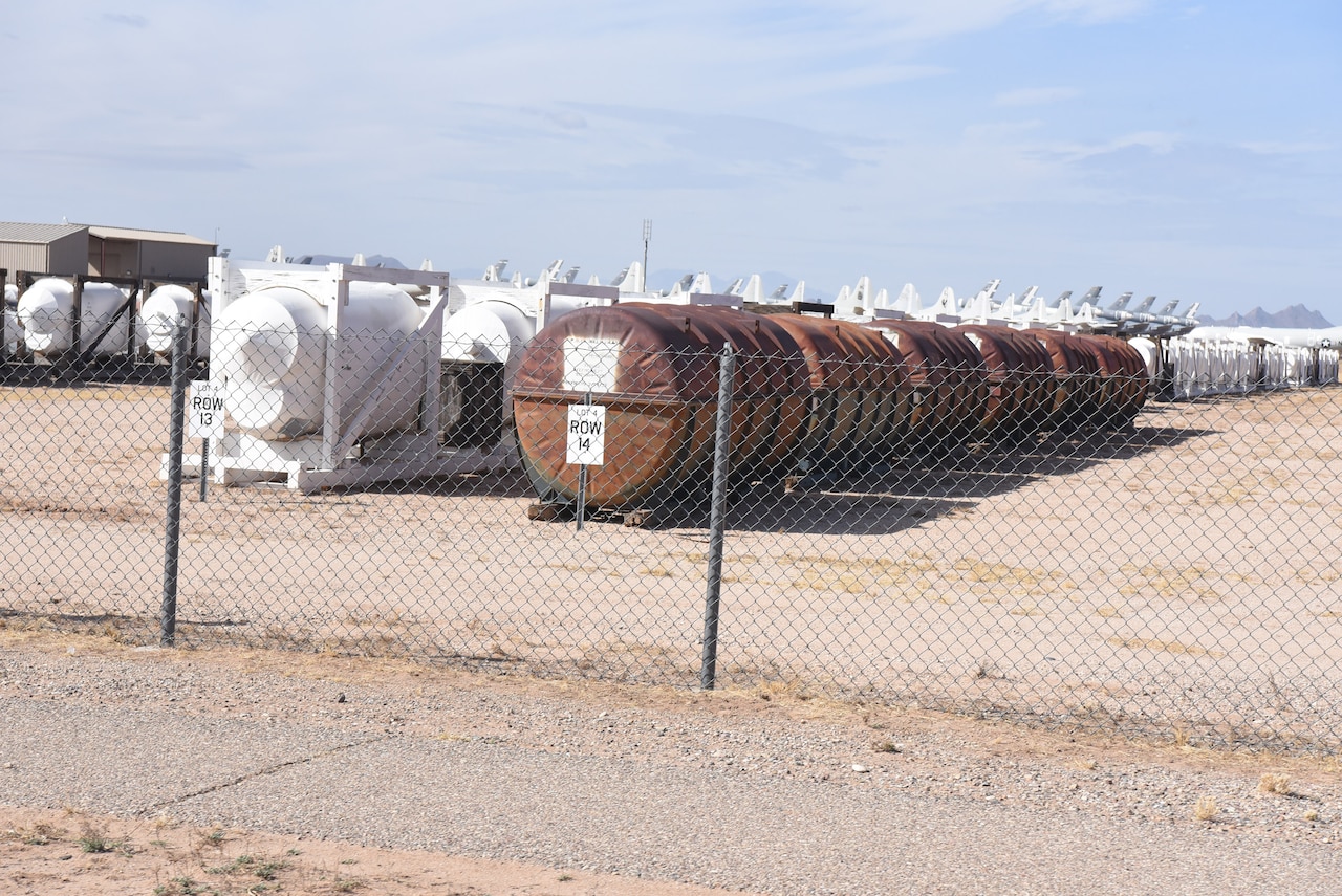 Aircraft parts in containers are lined up in rows in the desert behind a fence.