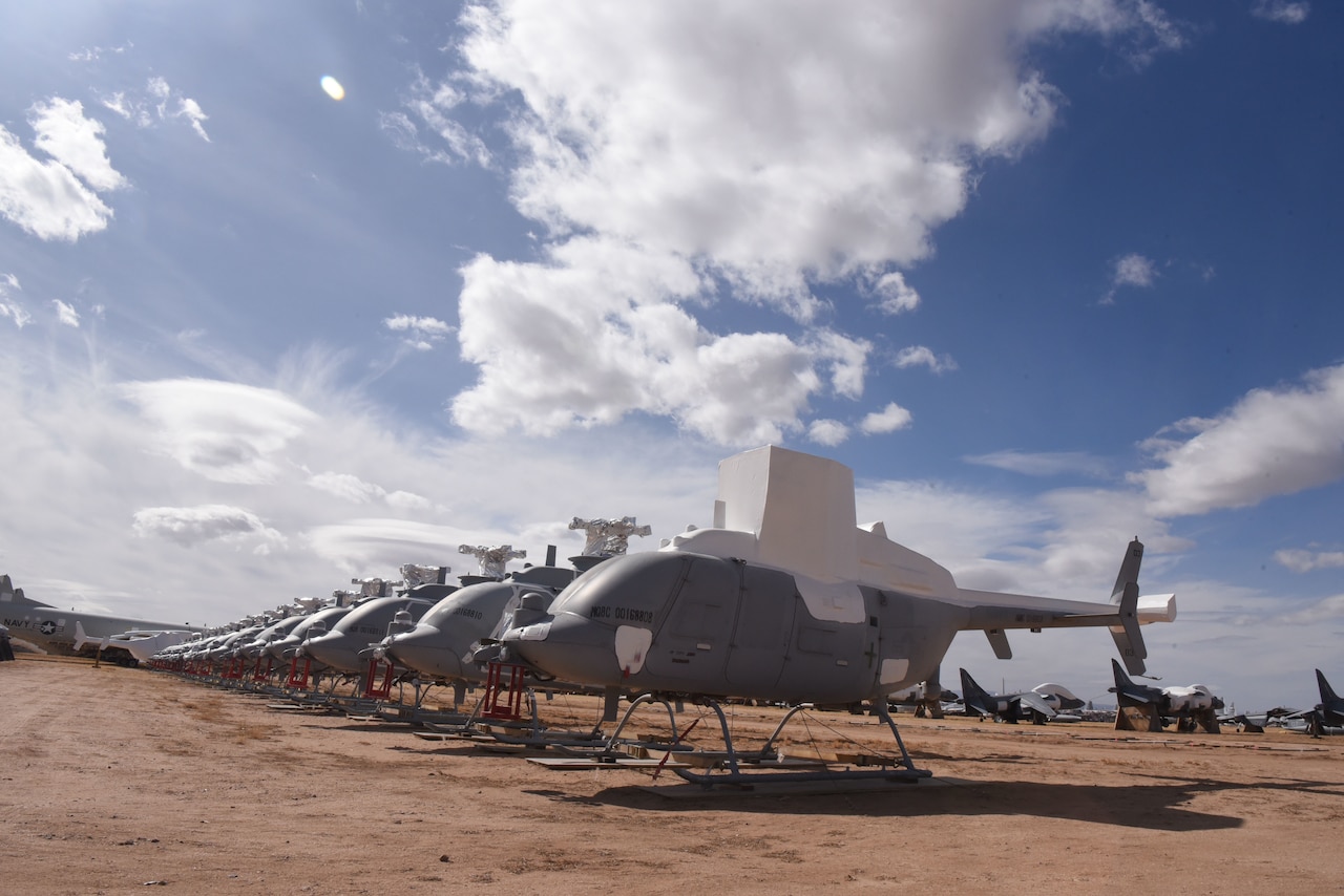 A row of drone helicopters without their propellers are stored in the desert.