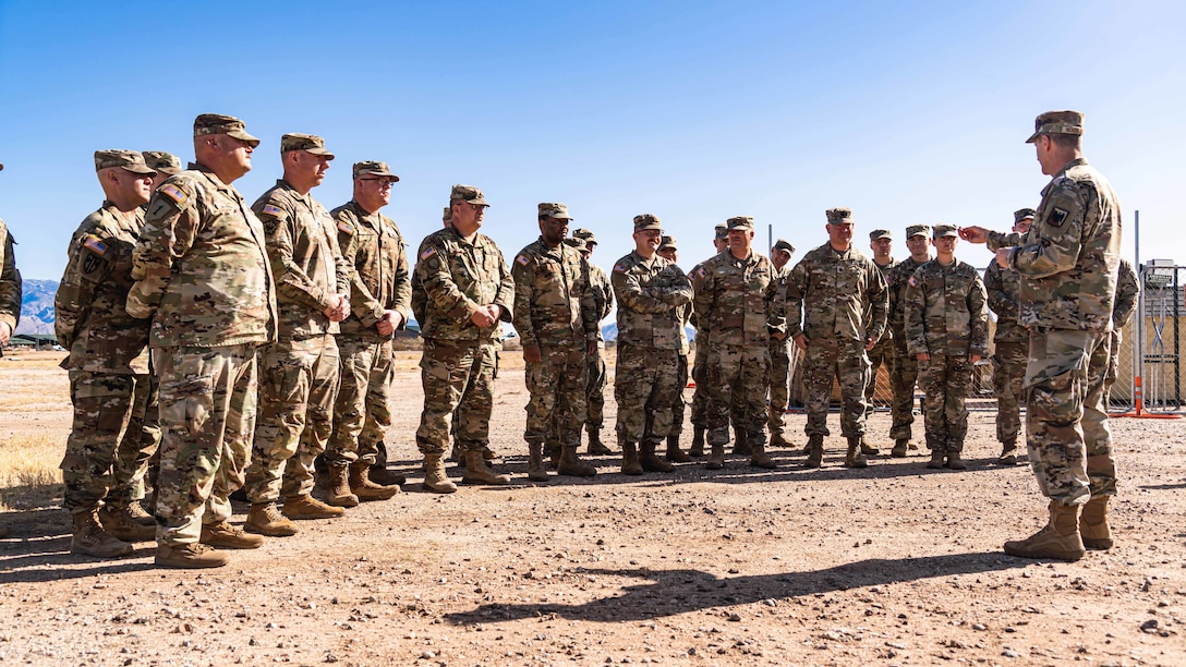 A group of service members form a semicircle around a single speaker in an open desert-like area.