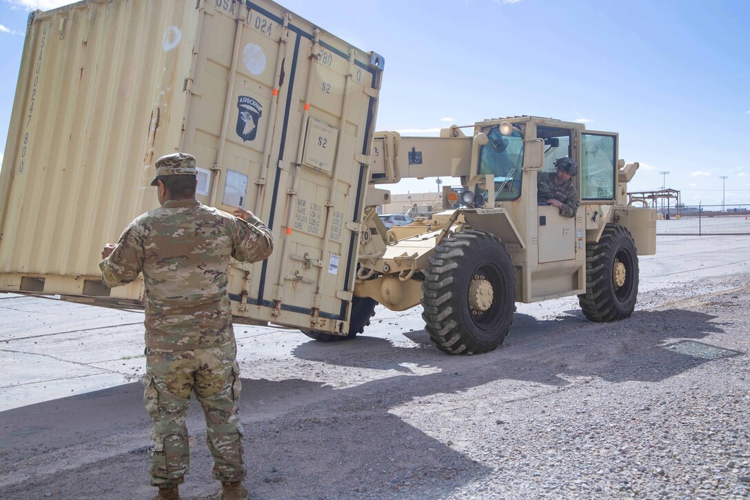 A soldier signals another operating a small vehicle to lower a large container to the ground.