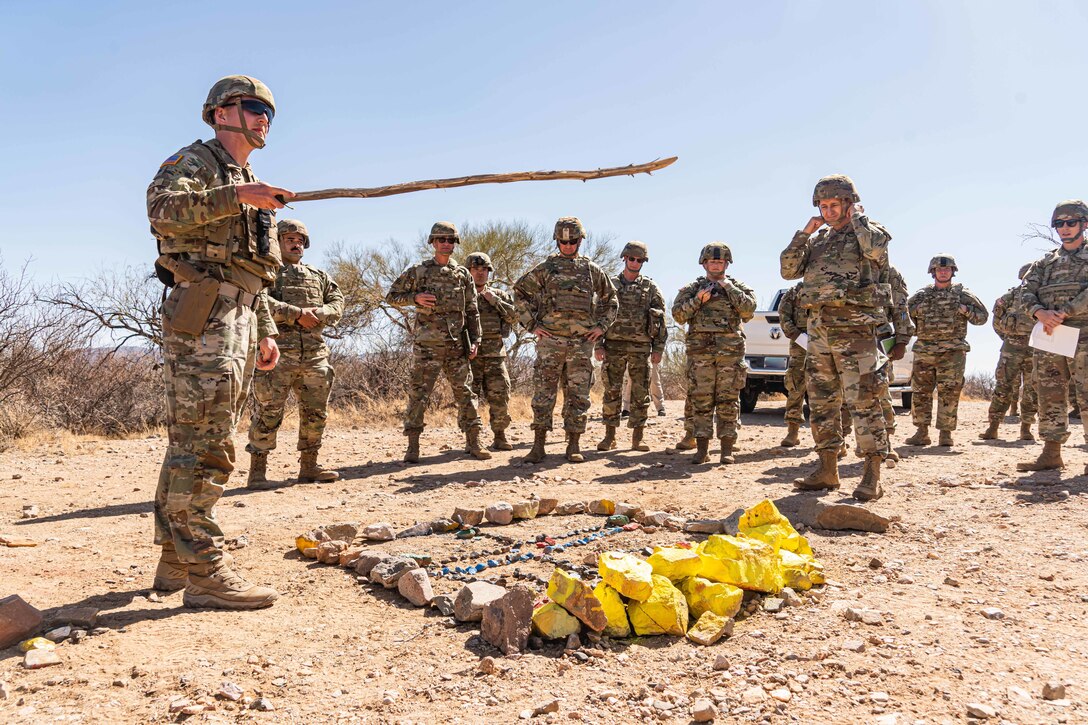 A soldier signals with a cane to a group of other service members around a demo pile on dry, desert-like ground.