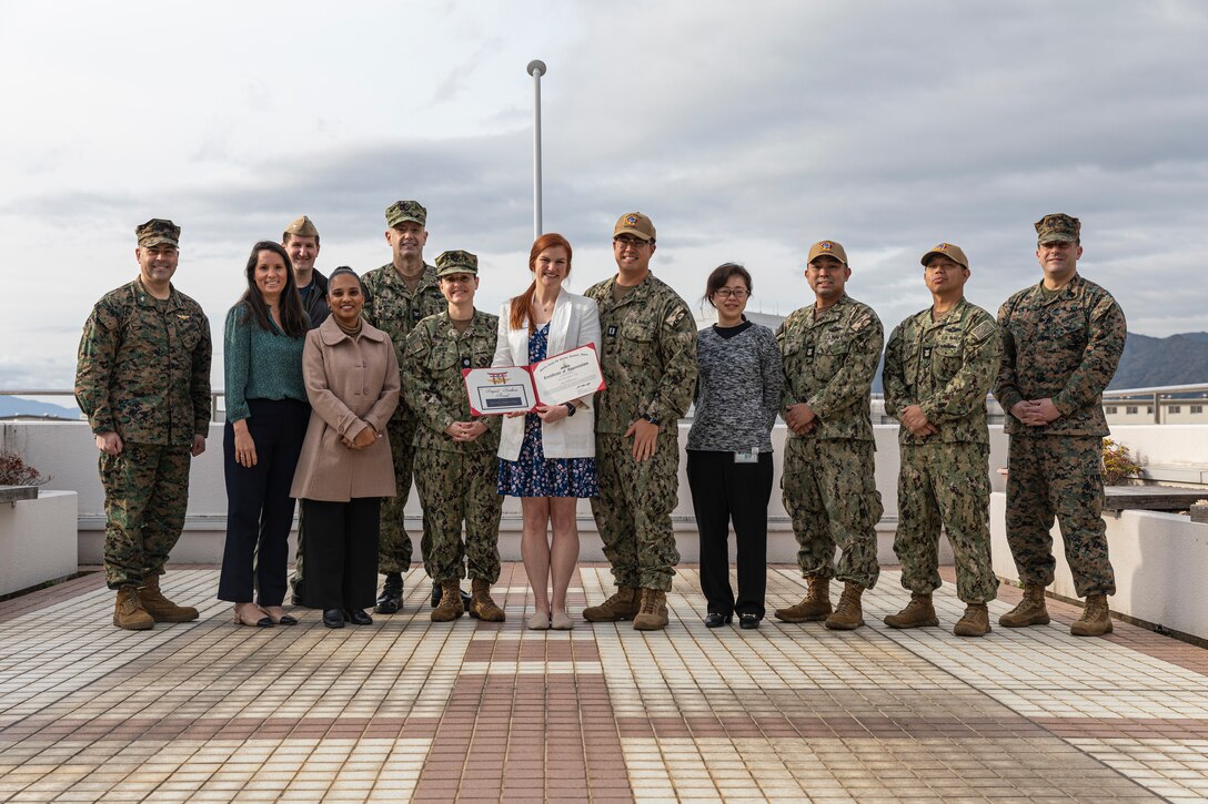 U.S. Marine Corps Col. Richard Rusnok Jr., left, the commanding officer of Marine Corps Air Station Iwakuni, and a Pennsylvania native, GySgt. Michael Moore, right, the station first sergeant of MCAS Iwakuni, and an Ohio native, Mary Kate Magdycz-Tong, a volunteer with the U.S. Navy ombudsman program, and members of the Branch Health Clinic pose for a group photo during the March 2025 Impact Iwakuni award ceremony at MCAS Iwakuni, Japan, March 6, 2025. The Impact Iwakuni award is presented monthly to recipients that make a significant positive impact on the community. The award went to Mary Kate Magdycz-Tong, who supported Sailors and their families by creating newsletters to inform them about events occurring on the installation. (U.S. Marine Corps photo by Cpl. Dahkareo Pritchett)