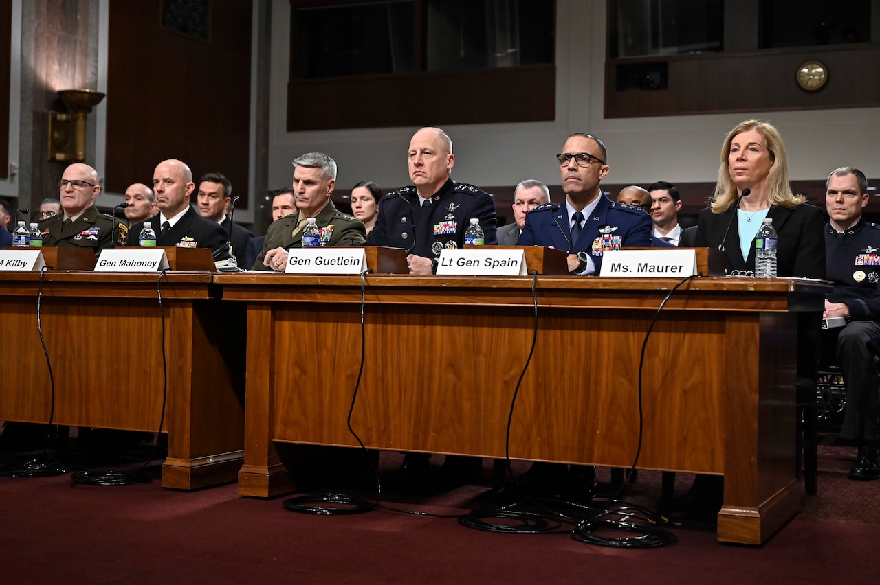 Military leaders and one civilian sit at a long table behind name placards and microphones