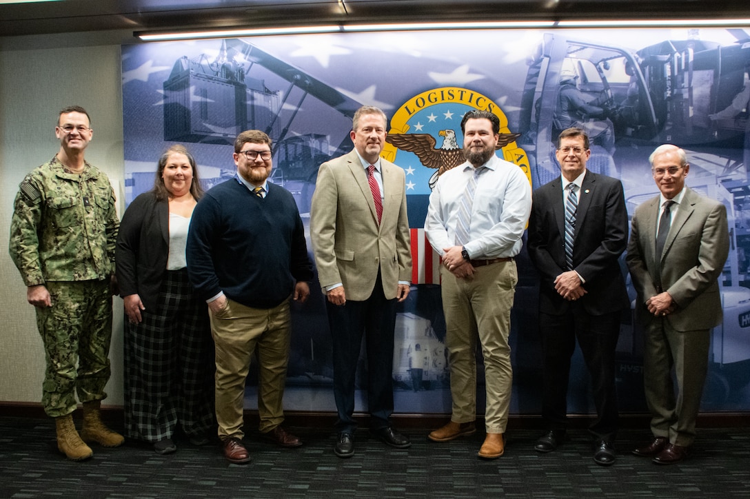 Six people stand in front of a wall with the Defense Logistics Agency logo on it.