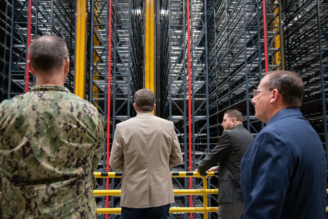 Four men stand in a warehouse looking up at a tall shelving/rack system.