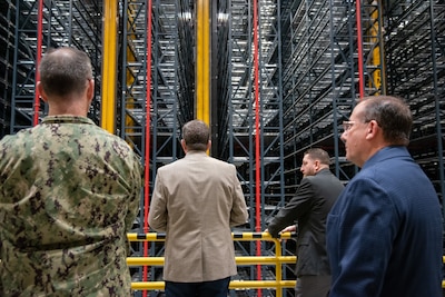 Four men stand in a warehouse looking up at a tall shelving/rack system.