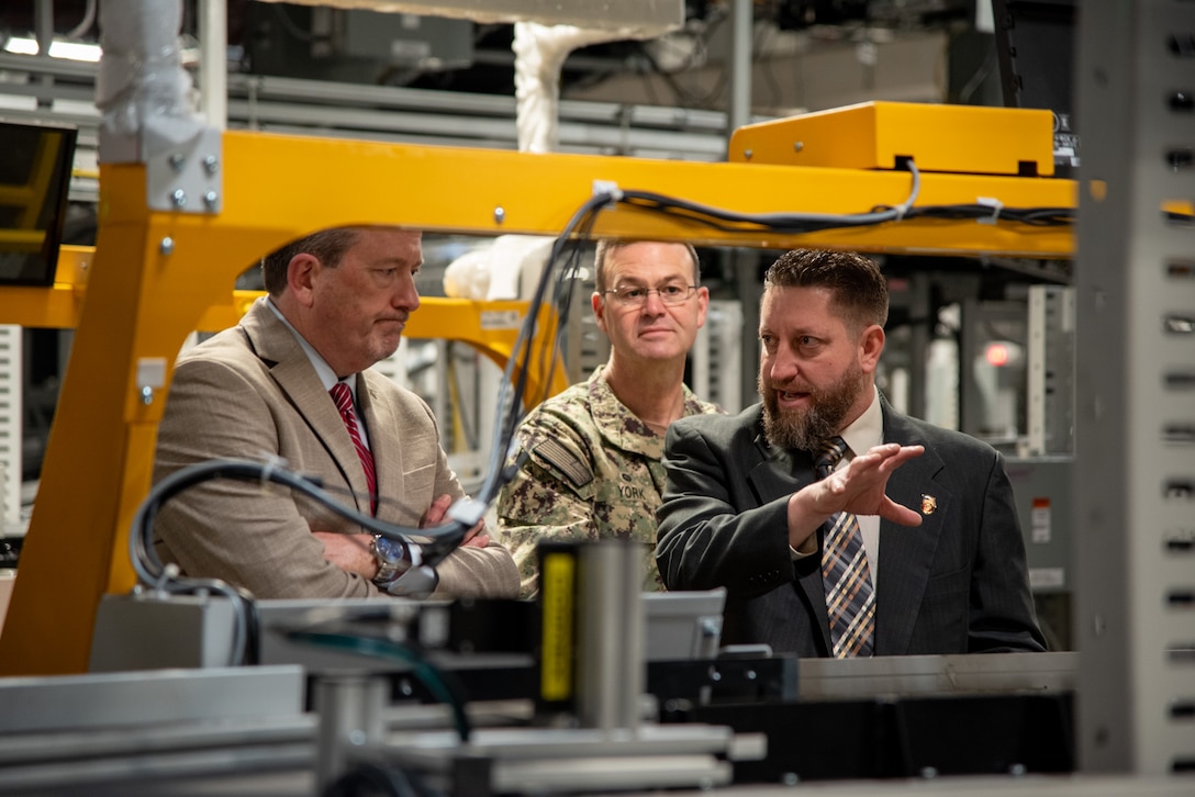 Three men stand in a warehouse behind a piece of machinery with the man on the right pointing to the machine.