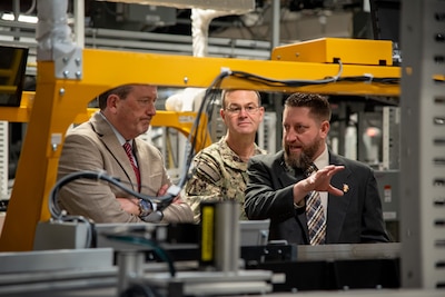 Three men stand in a warehouse behind a piece of machinery with the man on the right pointing to the machine.
