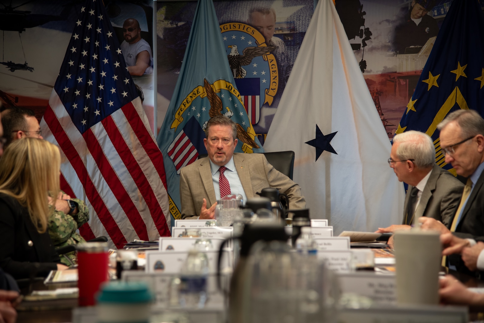 People sit around a large conference table looking toward a man seated at the head of the table with an American flag behind him.