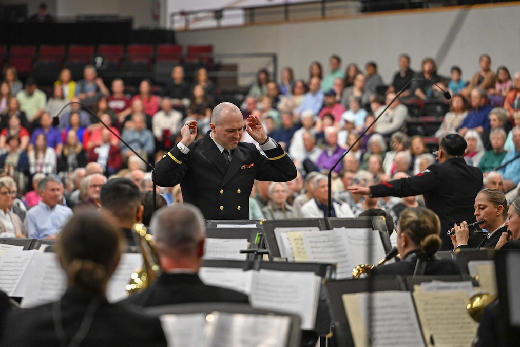 A sailor gestures while conducting a seated band in front of a seated audience.