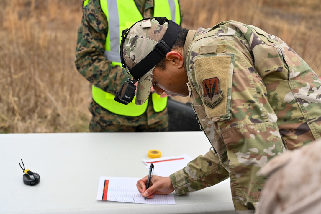 An Airman with a Go-Pro Camera on the brim of his hat bends over to write on a piece of paper his name and record his ruck sack weight prior to the start of the Norwegian Foot March.