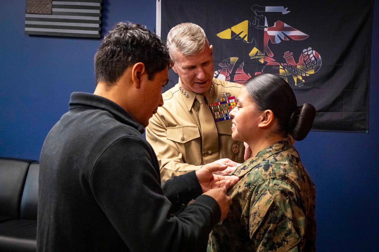 A Marine Corps officer pins an enlisted Marine with help from another person in civilian clothing against a dark Marine Corps flag on a blue wall.