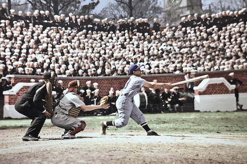 In a sideview, a batter hits a baseball at home plate of a packed baseball stadium in front of a catcher and an umpire