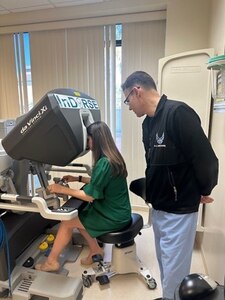 A serviceman in baby blue scrubs wearing a black hoodie with the Air Force logo on it stands over a woman in a green dress sitting down, operating machinery.